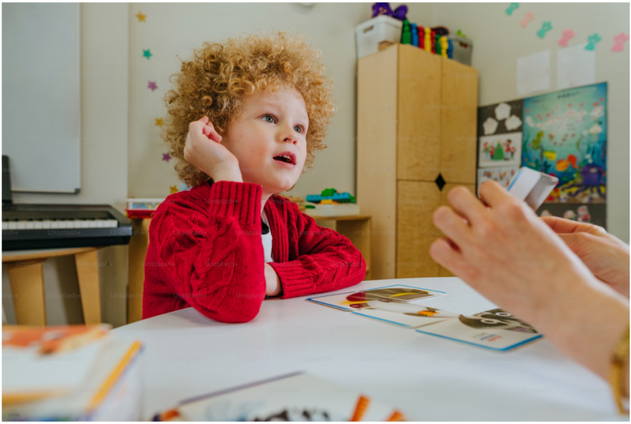 Two young children. A young curly brown haired girl is whispering into the ear of a boy with straight brown hair laughing.